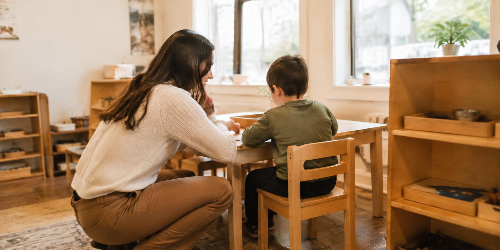 An educator kneeling at eye level beside a child at a classroom table